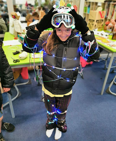 Student wearing a ski suit and wrapped in fairy lights with their hands holding a pair of goggles on their head. 