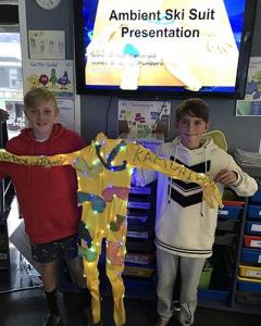 Two students holding up their light suit in front of a projected slide which reads, “Ambient ski suit presentation” . 