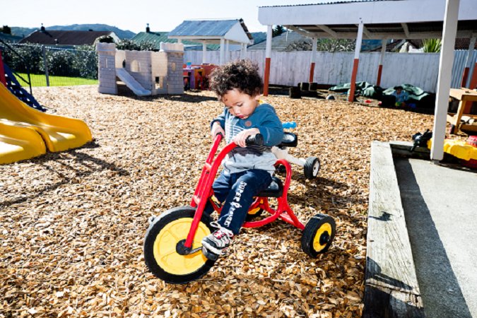 A child is riding a trike and looking at the pedals making the wheels go around.