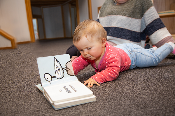 A baby is lying on the floor looking at a book and turning the pages themselves