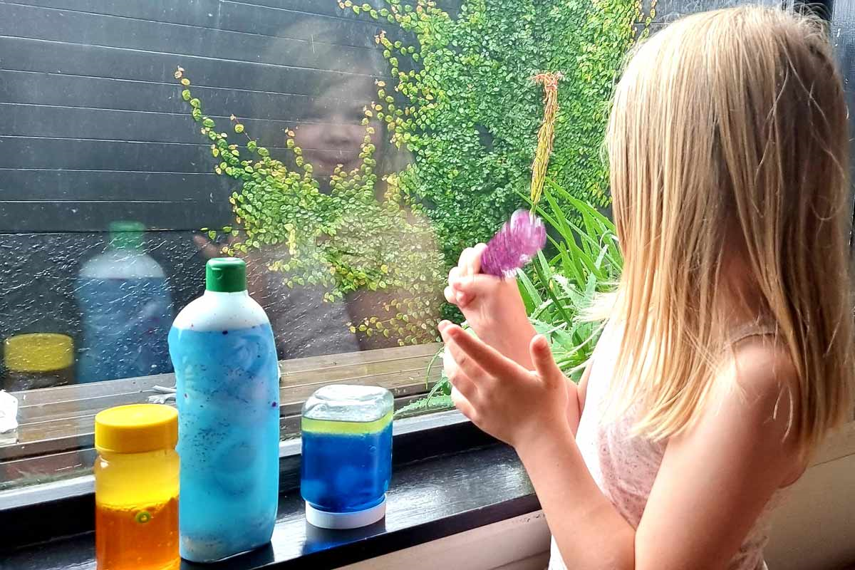 A child is playing with a sensory bottle by a window that shows her smiling reflection.