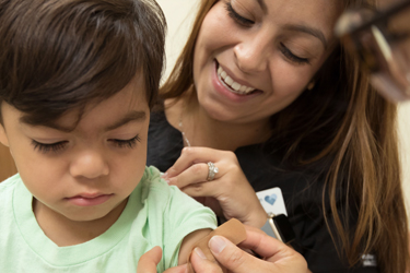 child with two adults. One adult is applying band-aid to child’s arm