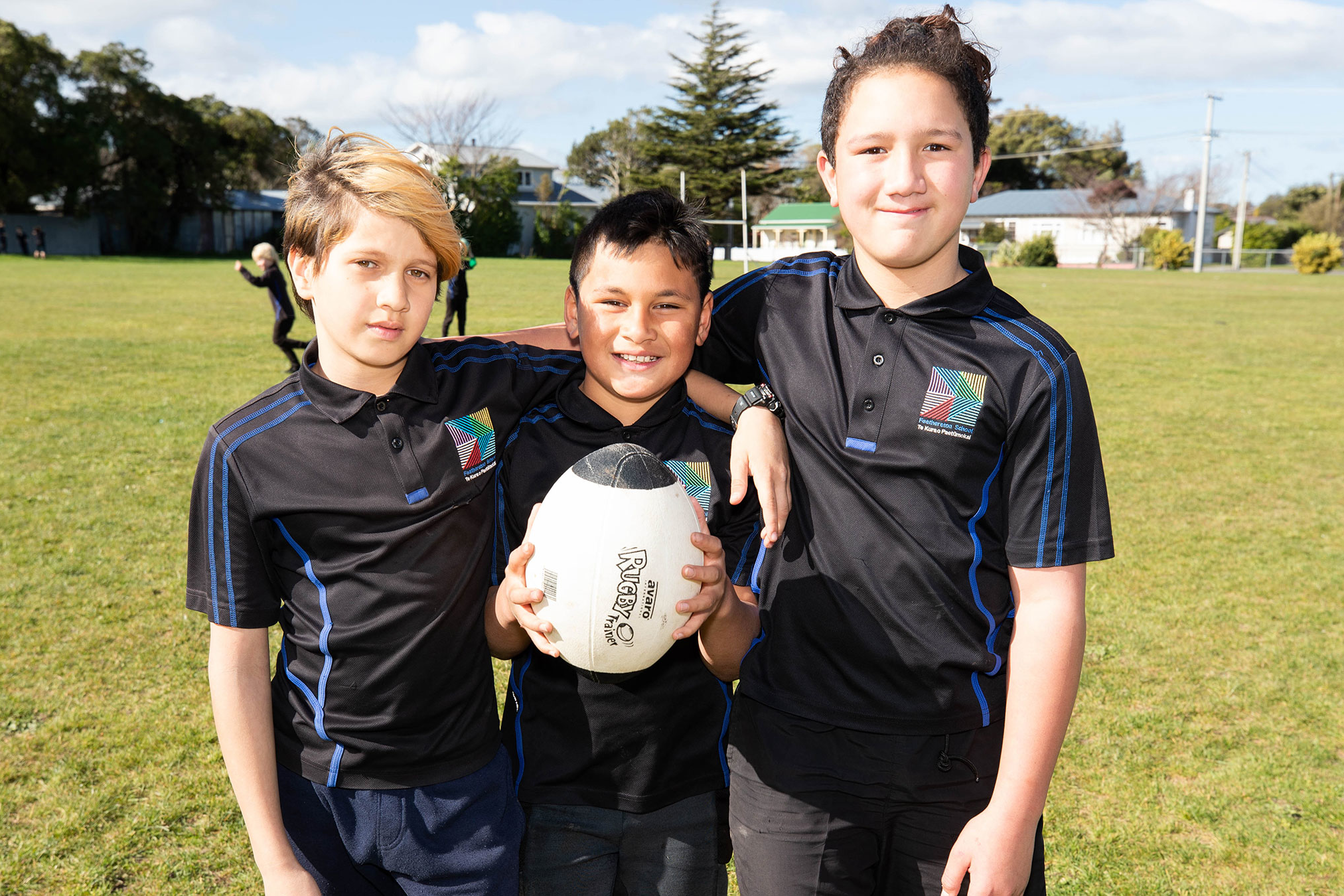 Three students in sport uniforms posing with a rugby ball and smiling.