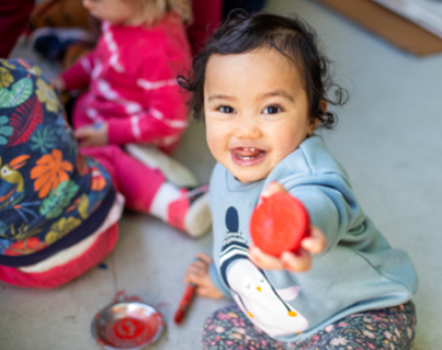 A toddler is smiling and handing an object to the person behind the camera.