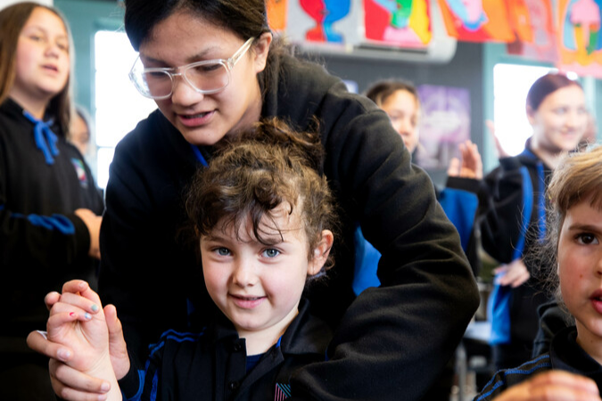 Older tamariki guides younger tamariki in teaching kapa haka movements.