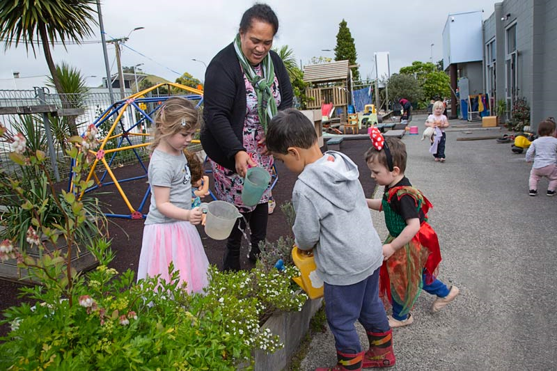 A kaiako and a group of children talk together while watering the garden.