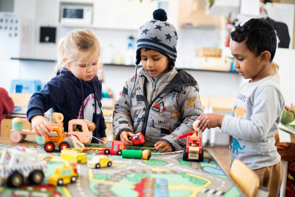 Tamariki play together with vehicles on a mat, a person shaped toy lies down.