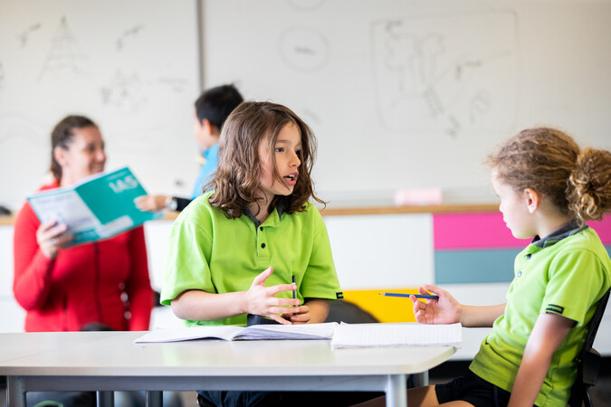 Two young students sitting at a table speaking to each other.