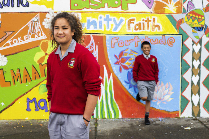 Two students stand in front of a Tongan art mural smiling at the camera.