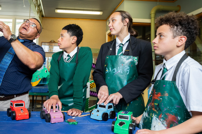 Students listen to their teacher as he speaks, all holding toy cars wearing aprons.