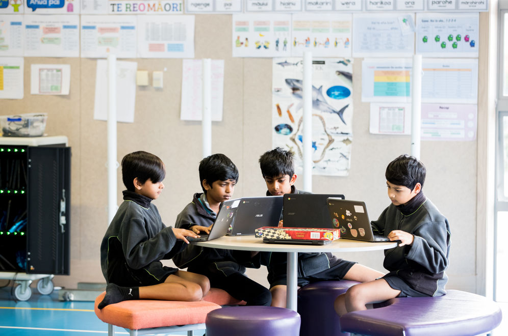 Four young ākonga sit around a circular table working on laptops.