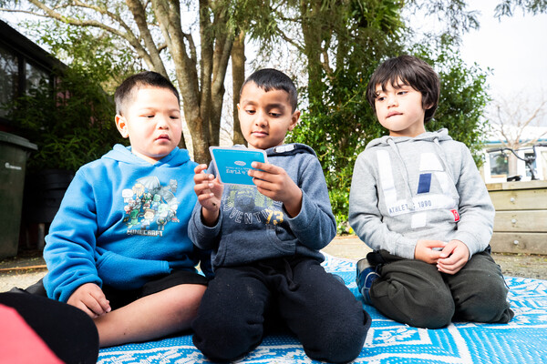 Three tamariki sit on a mat outside reading together.