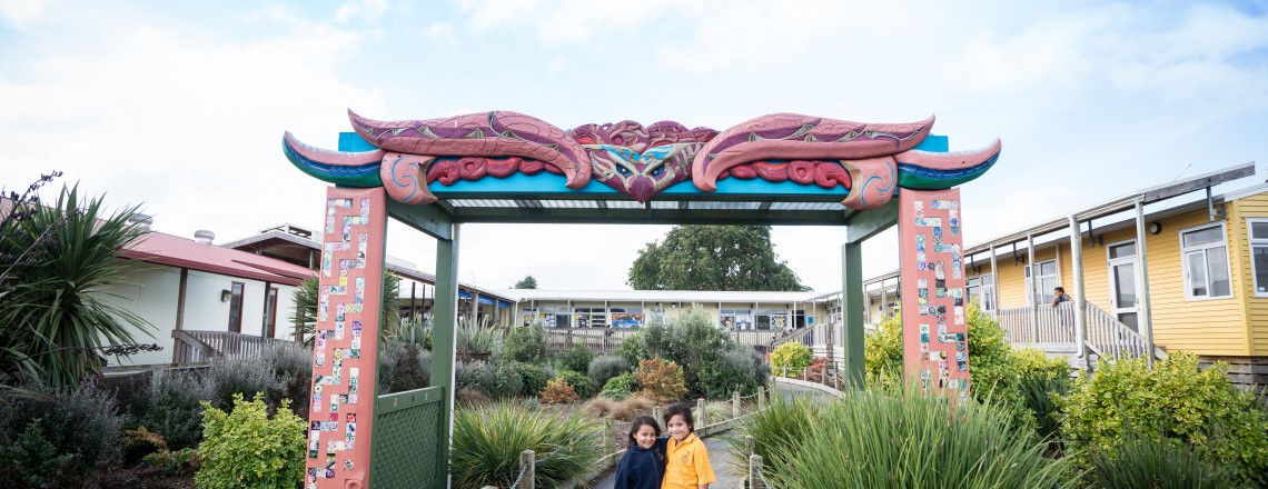 Two young tamariki stand smiling at the camera outside a school entrance. 