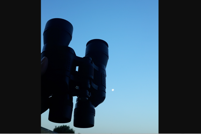 Binoculars silhouetted against a blue sky with a full moon.
