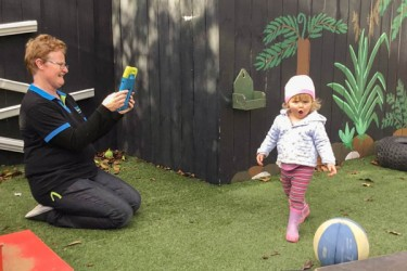 A teacher kneeling on the ground filming a child playing with a ball.