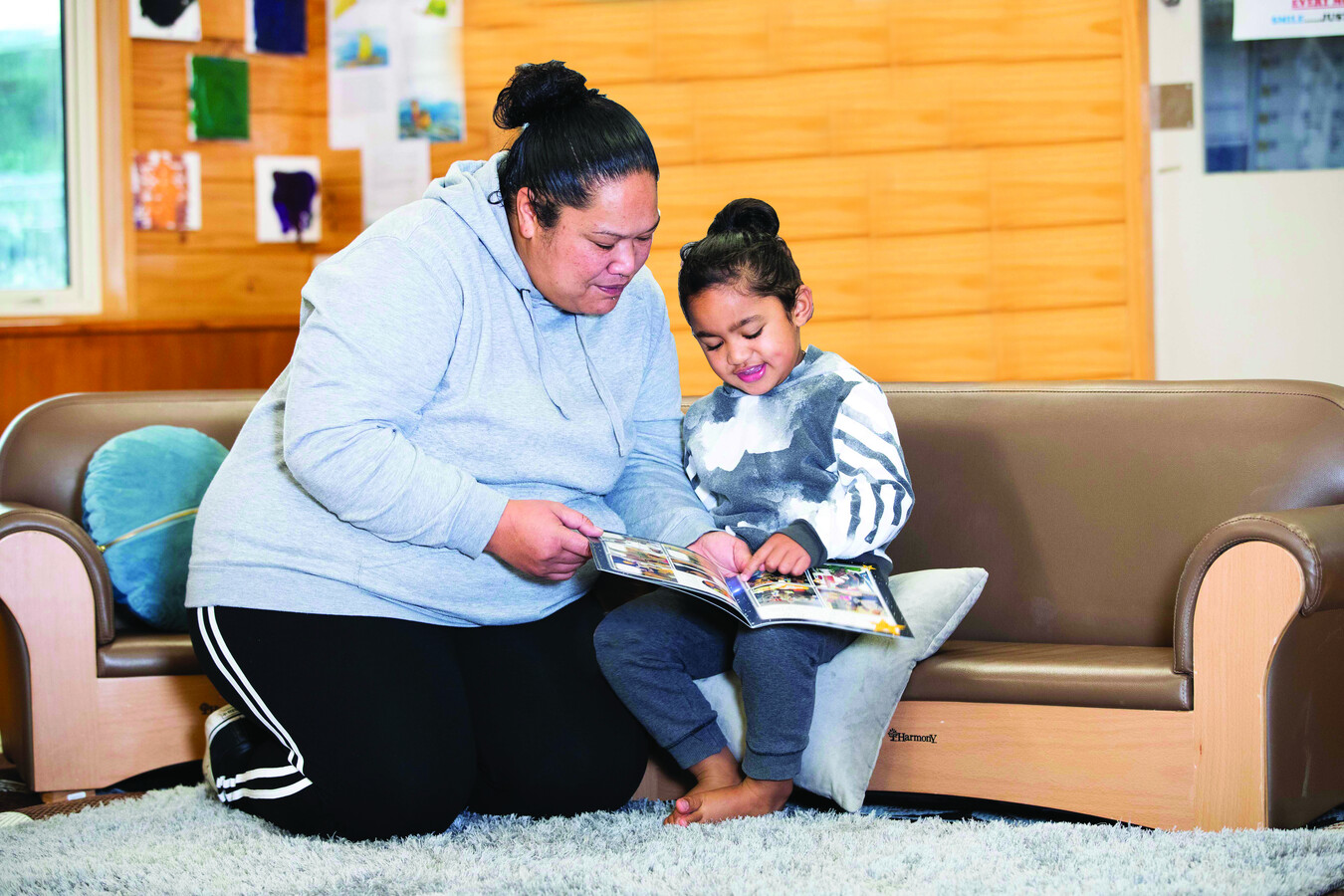 A Samoan mother and her young child reading a picture book together on the couch.