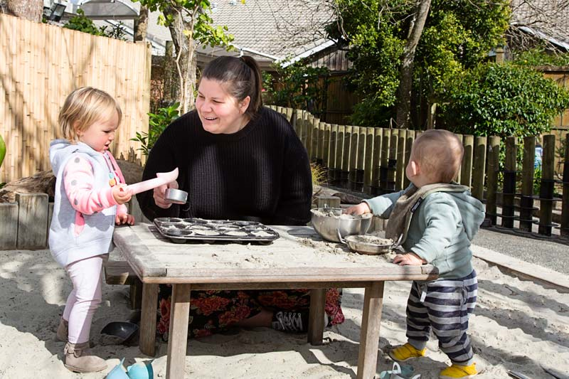 A kaiako talks with two tamariki while playing with bowls and sand