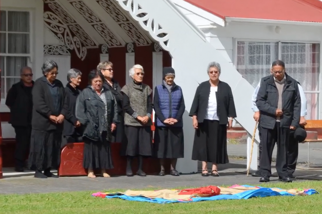 A photograph of an iwi standing outside the marae participating in a traditional ceremony. 