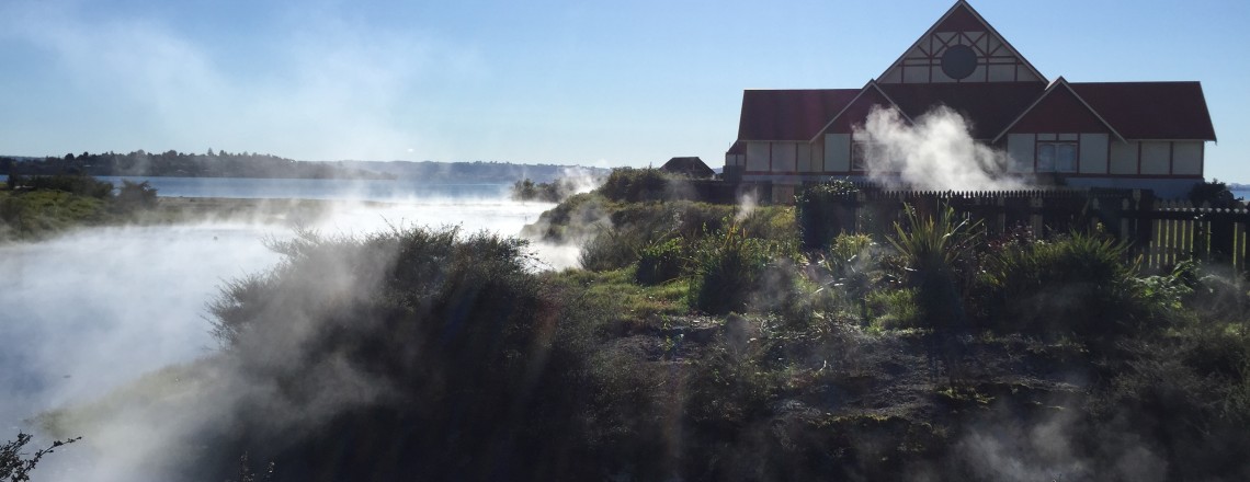Photograph of steaming hot springs with a house in the background. 