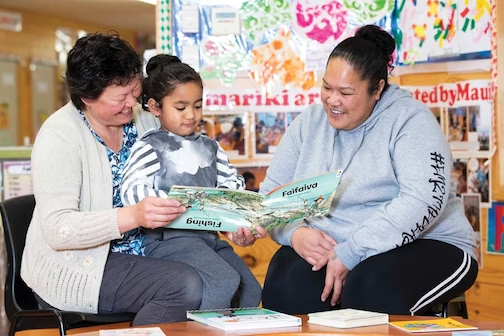 A teacher sitting with a child on her mum's lap reading a book.