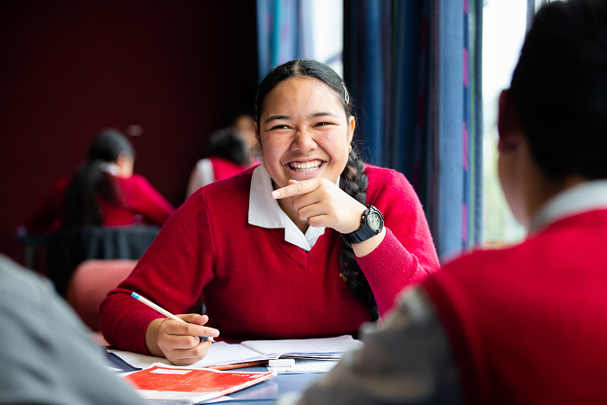 Ākonga sitting in classroom smiling at camera as they write in their workbook.