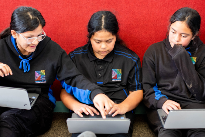 Three young girls in school uniform sitting on the floor with laptops on their stretched out legs.