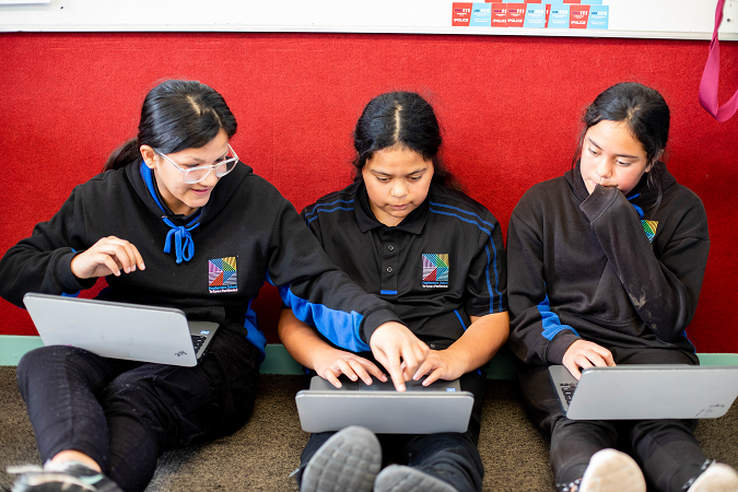 Three girls sitting down in a classroom working on their laptops.