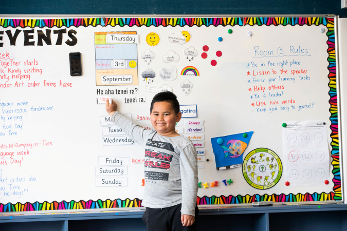 Tamariki stands in front of the classroom whiteboard smiling. 