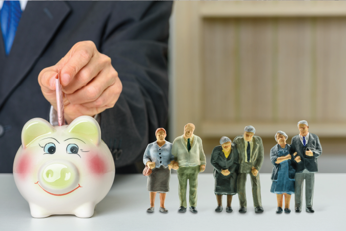 A hand putting money into a piggy bank, a series of figurines of elderly couples stand next to the piggy. 