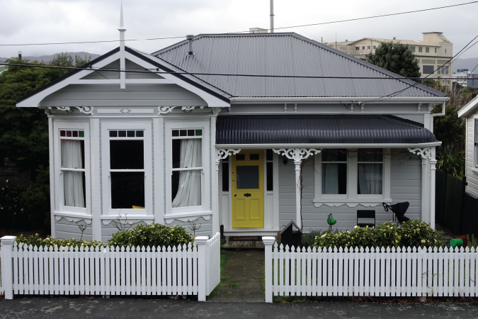 Grey villa with grey roof a yellow door and white picket fence.