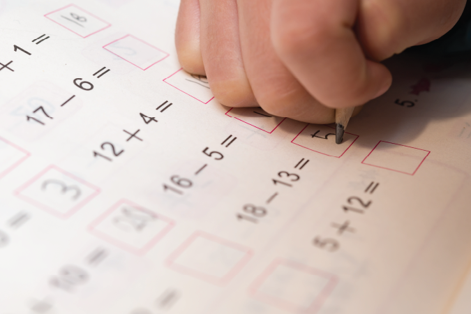 A student's hand holding a pencil, filling in the answers to a list of maths equations printed on paper.