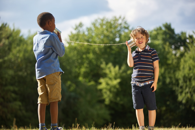 Young boys standing outside, one speaking into a can connected by string, the other holding the can to his ear listening.