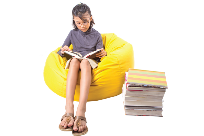 A girl sitting on a yellow bean bag reading a book with a pile of books on the side.