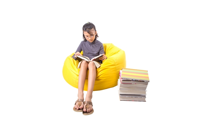 A girl sitting on a yellow bean bag reading a book with a pile of books on the side.