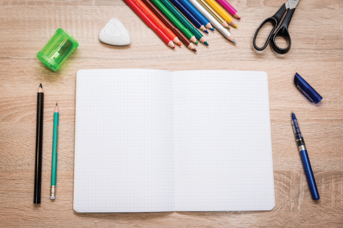 Open notebook surrounded by other stationary; pens, pencils, scissors, eraser, pencil sharpner. 