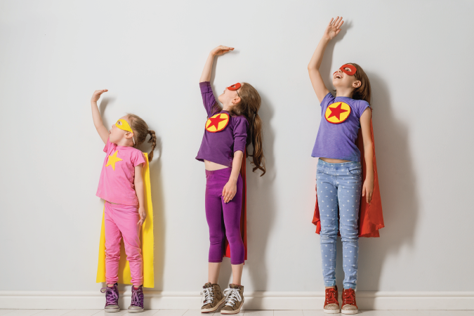 Three children in superhero costumes with their left arm stretched above them.