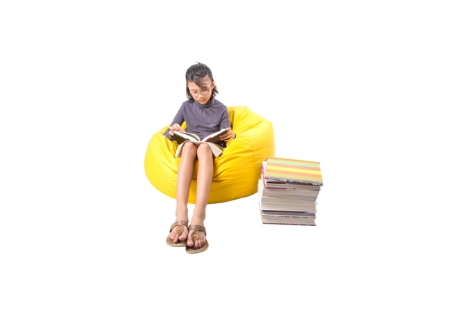 A girl sitting on a yellow bean bag reading a book with a pile of books on the side.