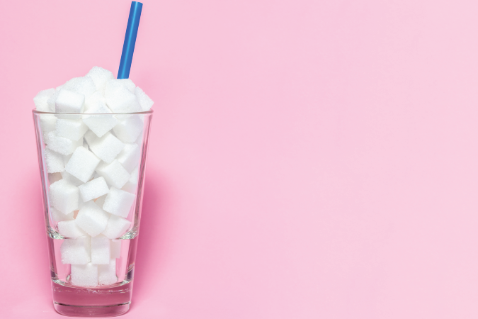 A glass filled to the top with sugar cubes, it also has a blue straw and is quarter filled with water on a pink background.