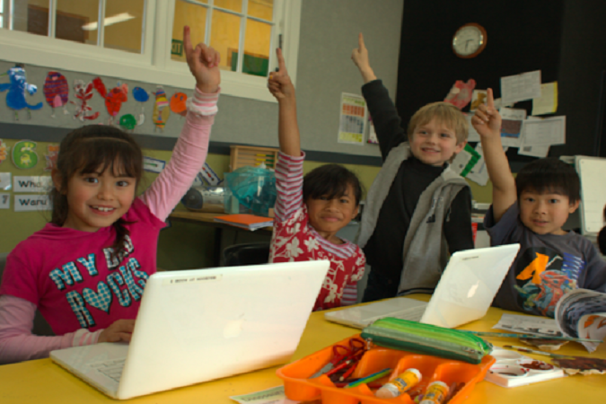 Students are at a table with devices with their hands up.