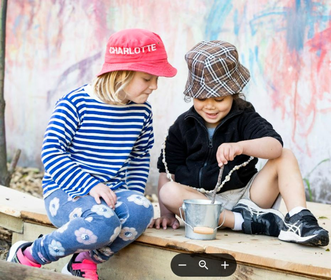 Outside two young children sit close together looking curiously into a small metal bucket. One child is stirring something in the bucket with a stick while the other child watches.