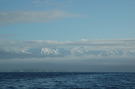 A body of water in front, a mountain range and clouds in the background. The tops of the mountains are covered in snow.