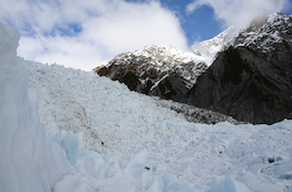 A glacier, with mountains behind it. There is snow on the tops of the mountains.