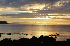 This is a sunset. The sun is sitting above an island on the horizon. There is a calm sea in the foreground with a rocky outcrop.