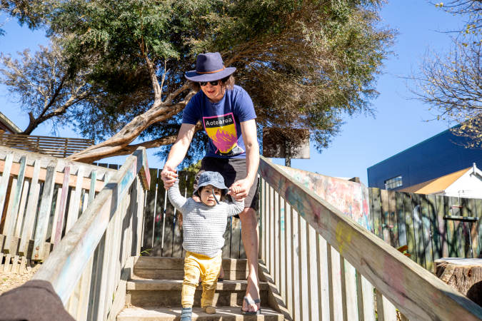 Toddler walking down steps outside with kaiako. Kaiako is holding the child's hands to support them walking.