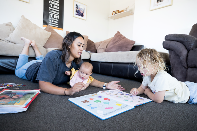 Infant and child on the floor with an adult in a home discussing a book with shapes in it.