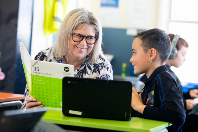 A student sitting at a desk with an open computer speaking with a teacher who is holding an open exercise book.