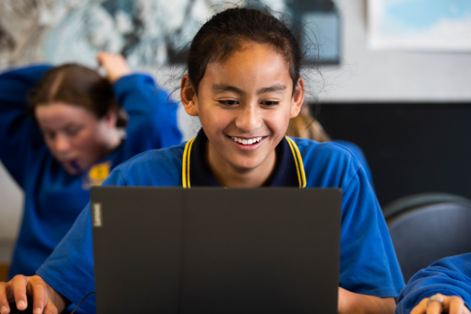 A student sitting at a classroom table, smiling while looking at a computer screen.