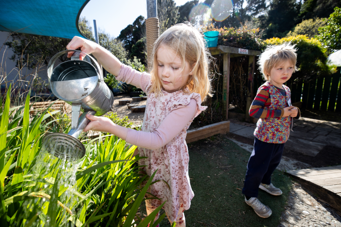 Two children outside. One child is watering a plant with a watering can.