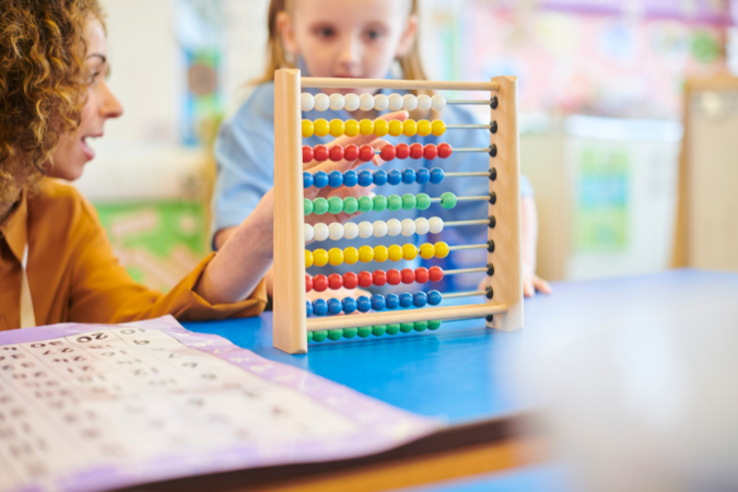 Teacher and student sit together in the classroom counting numbers on an abacus. 