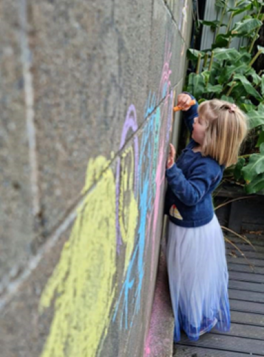 A young girl draws on an outside brick wall with chalk.